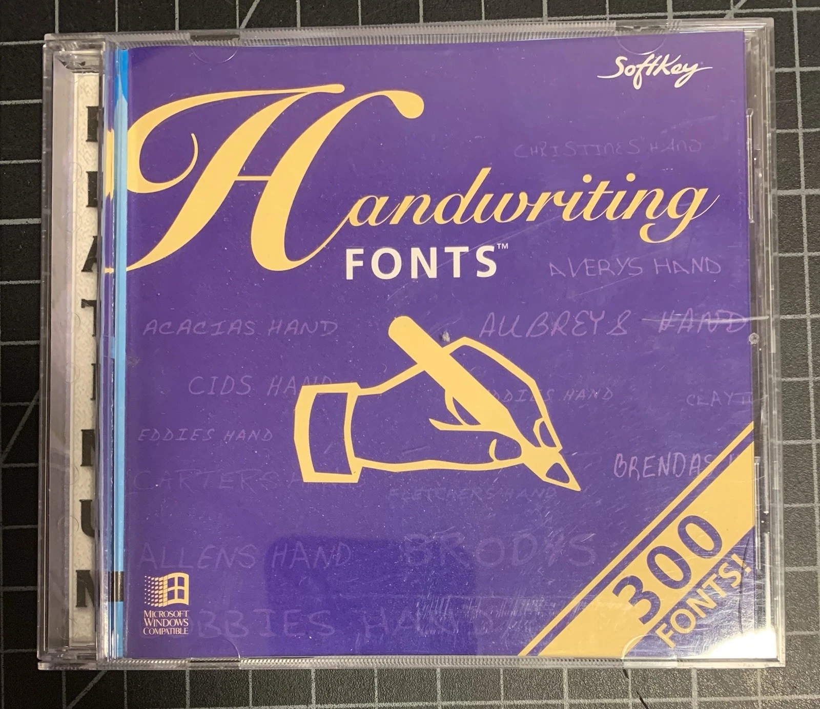 The Handwriting Fonts CD jewel case: purple cover with a hand holding a pen, the names of its handwriting fonts ghosted across the design, and a banner promising 300 fonts.